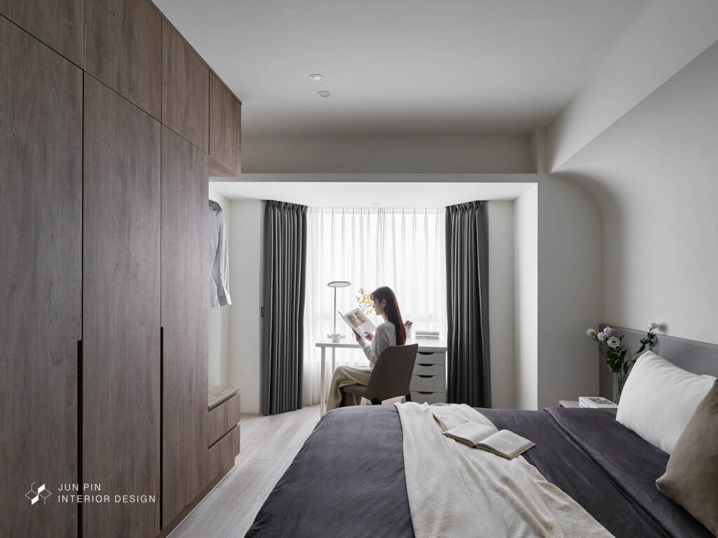 臥室整體以白色及淺色木頭為主， 讓臥室散發溫暖柔和的質感， 透過弧形的設計修飾床頭壓樑的結構， 並在系統櫃靠近門的那一側設置汙衣吊衣區， 在視覺上也有不會過於單一壓迫的效果。
