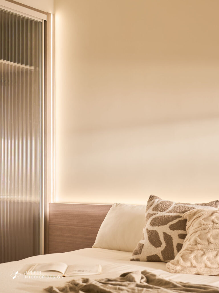 主臥以隱藏門連接客廳，延續整體立面整潔感。 床頭背板使用淺木飾面，搭配鋁條燈光帶，營造出柔和的光影氛圍。