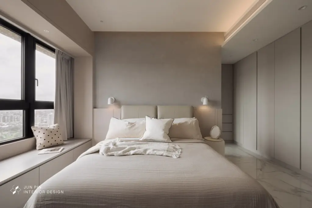 床頭牆面使用篇灰色的特殊漆作為床頭裝飾，省去過多繁雜的線條，利用本身就有層次的特殊漆帶出空間質感。左側窗邊設置了窗邊臥榻，上方的大量也用相同的奶茶色包覆，讓大樑和臥榻形成框景，弱化了大樑的視覺，同時也為空間創造端景。